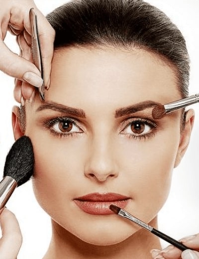 Beautiful woman looks forward as multiple hands with makeup brushes touch her face