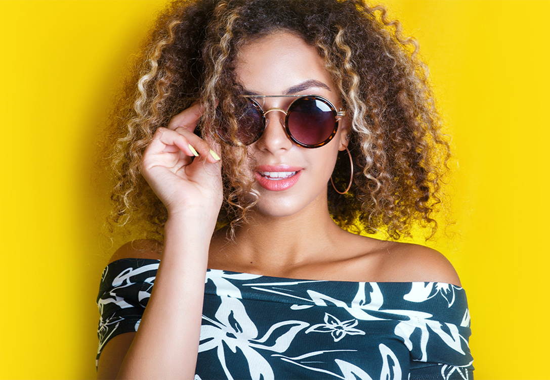Curly hair woman wearing off-shoulder dress touches round sunglasses on her face