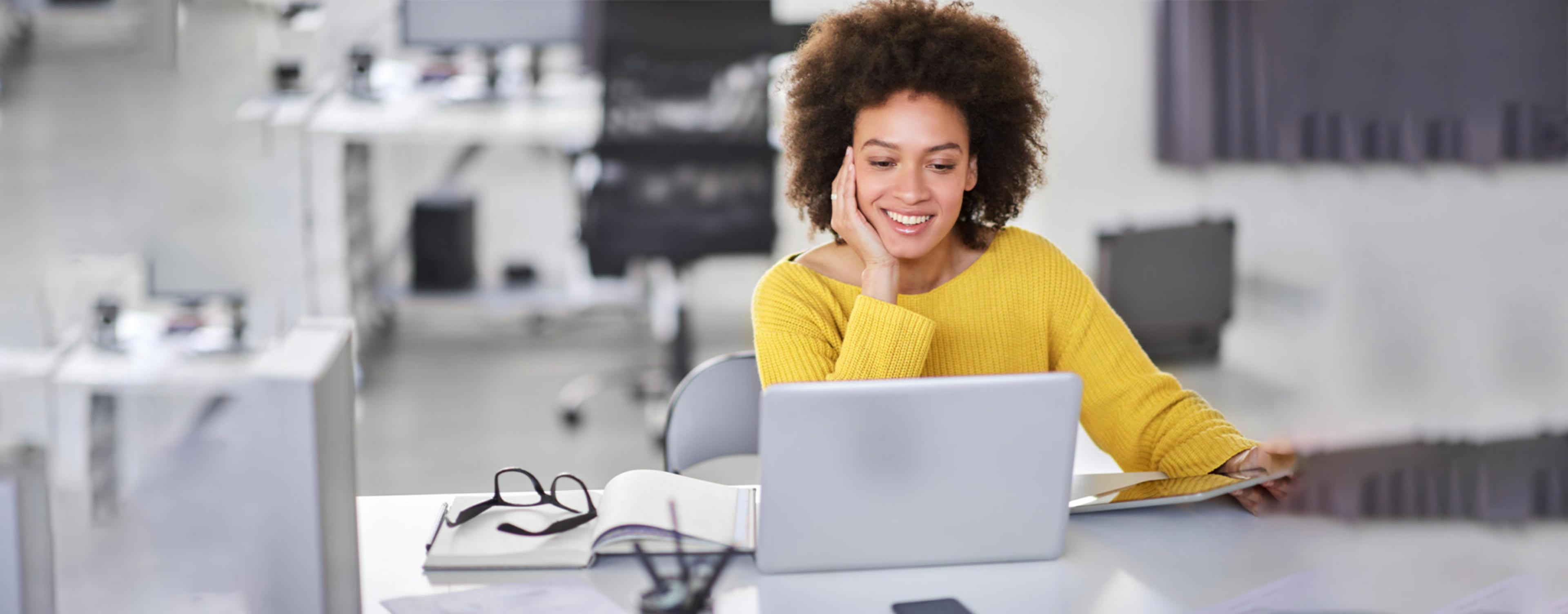 Beautiful woman wearing yellow sweater sits at office desk smiling at her computer