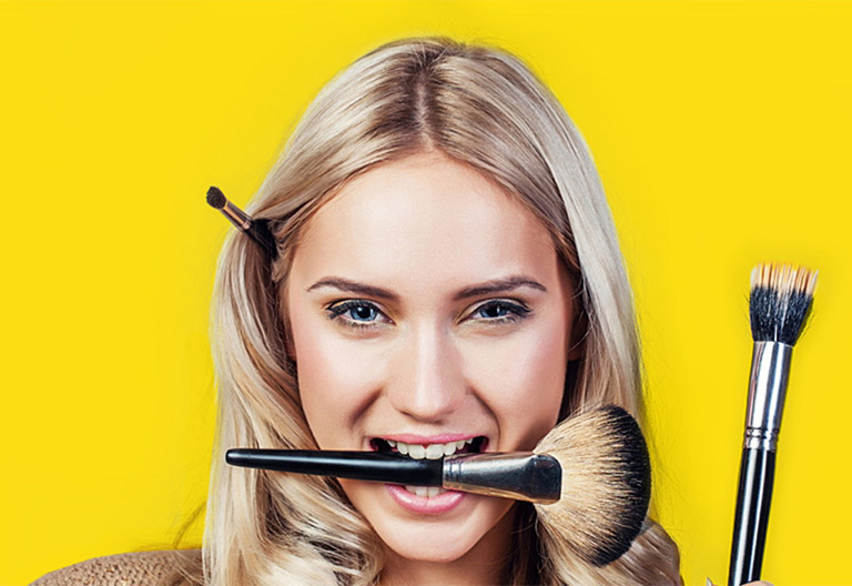 Mobile salon professional woman with blonde hair poses while holding makeup brushes in her mouth, hand, and behind her ear
